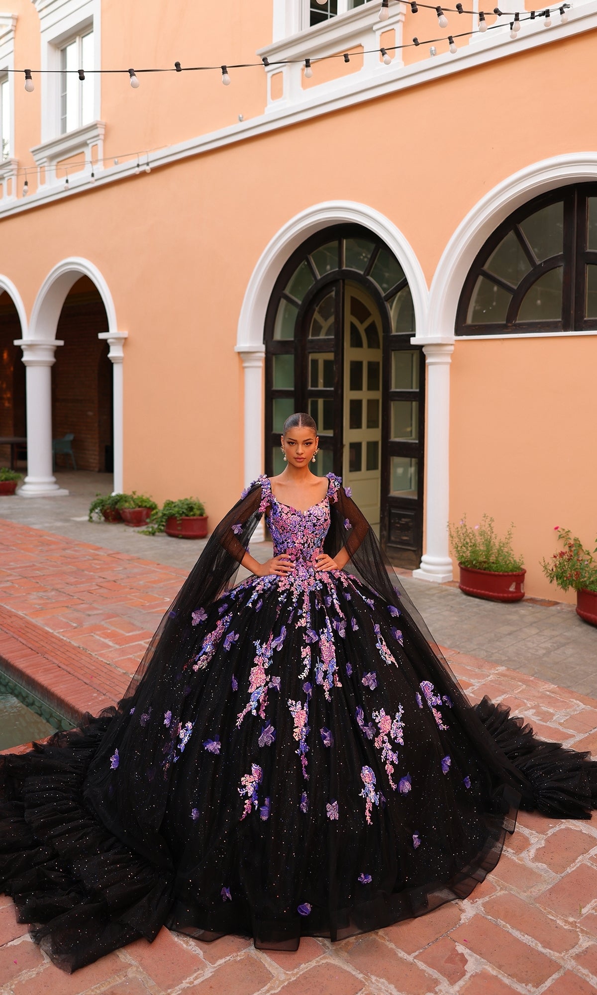 Outdoors before an arched building, a person wears Amarra's Quinceanera Dress 54318 featuring purple and pink floral details, black tulle accents, and elegant floral embroidery.
